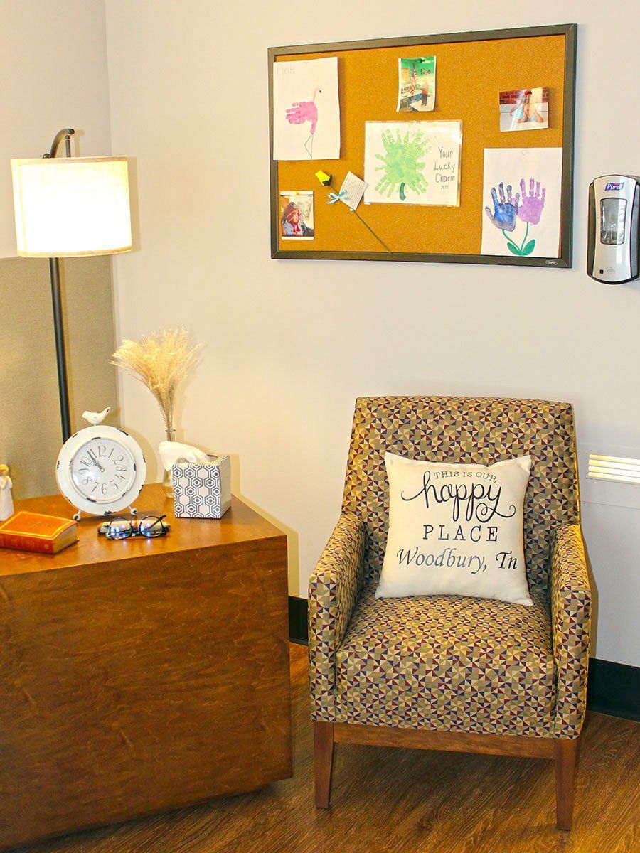 Cozy office corner: armchair with patterned fabric, bulletin board with artwork, desk with lamp and clock.