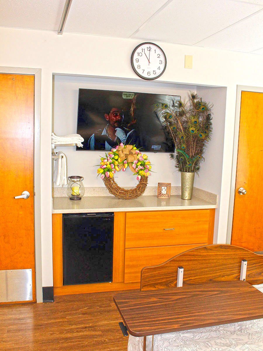 Built-in entertainment center in a room. Includes TV, mini-fridge, clock, wreath, and peacock feathers.