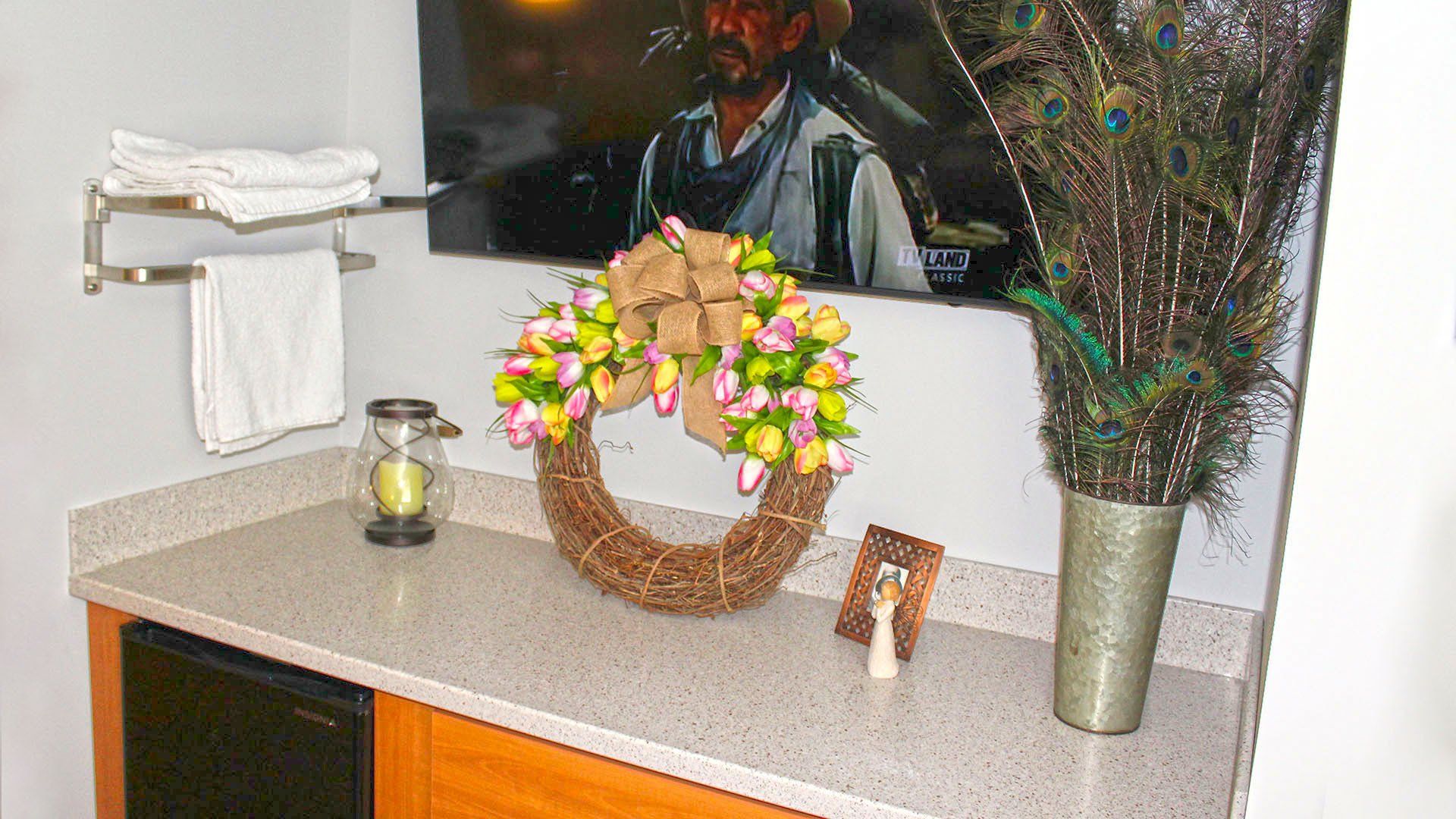 A hotel room counter with a floral wreath, peacock feathers, and decor. Towels and artwork are above.