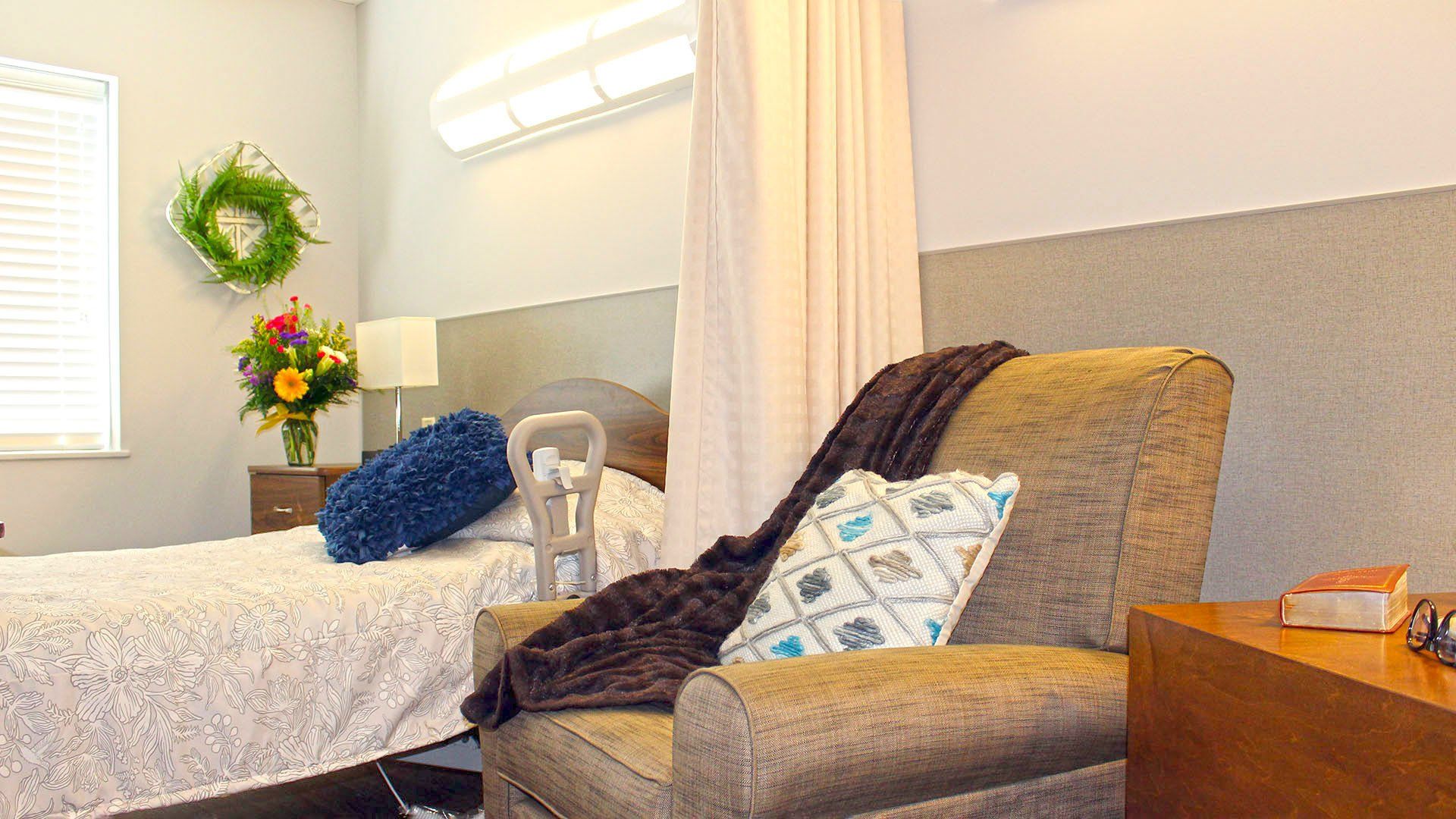 Bedroom with bed, floral arrangements, armchair, and neutral-colored walls. A curtain is visible near the bed.