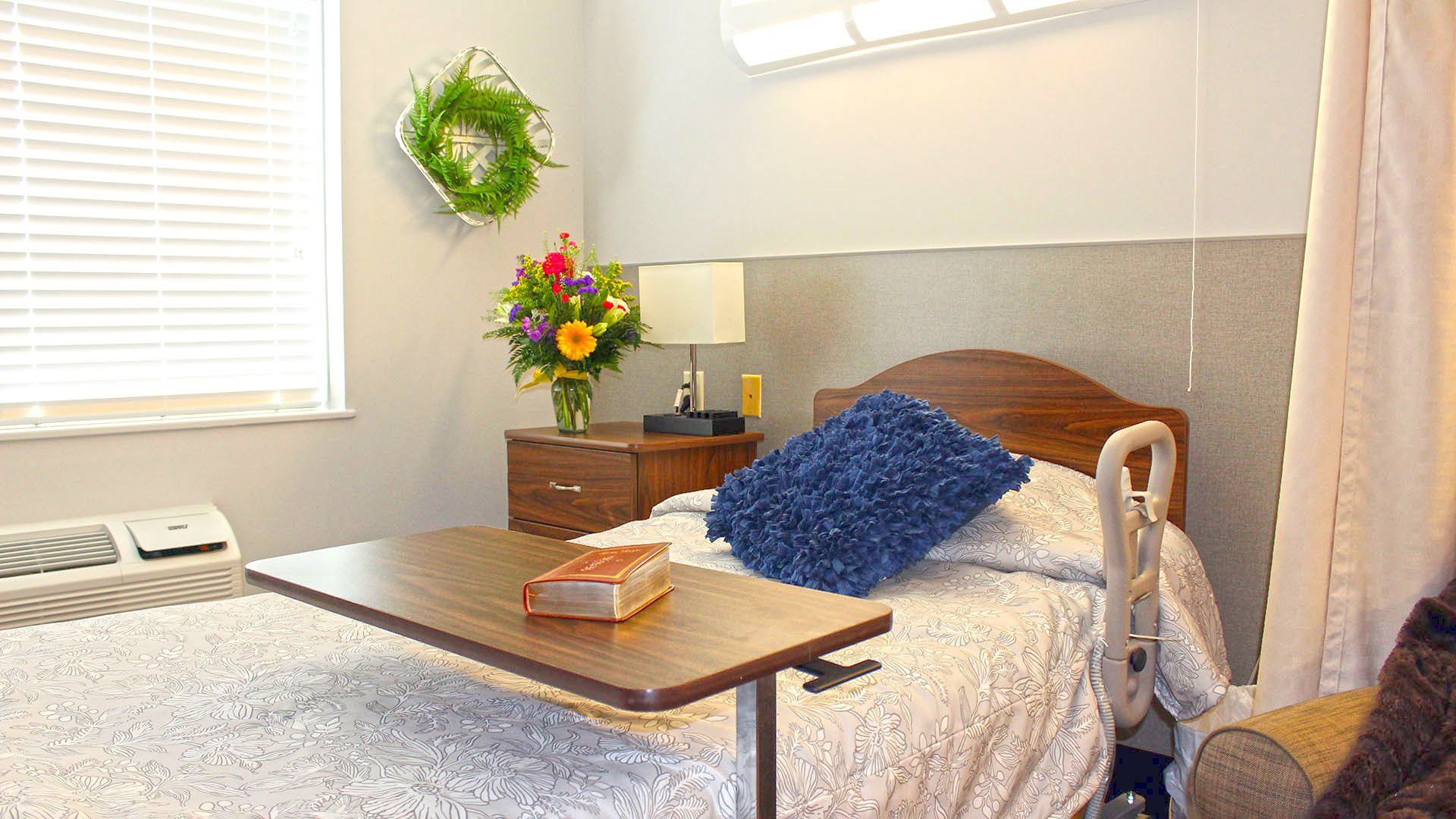 Cozy hospital room with bed, table, nightstand with flowers, and window with blinds.