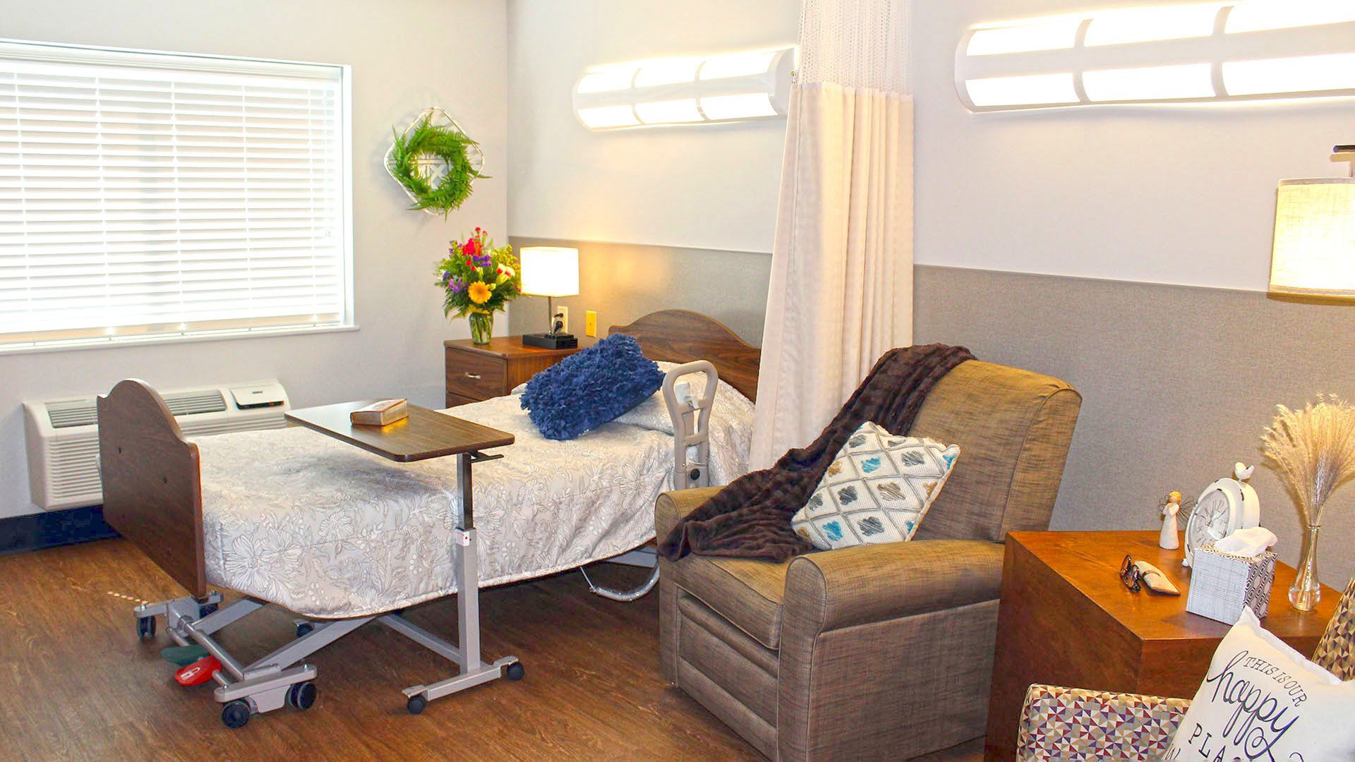 Hospital room interior with a bed, recliner, bedside table, and window.