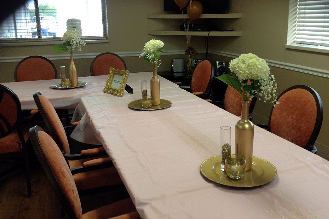 A long table set for a gathering, with pink tablecloth, flowers in golden vases, and chairs.
