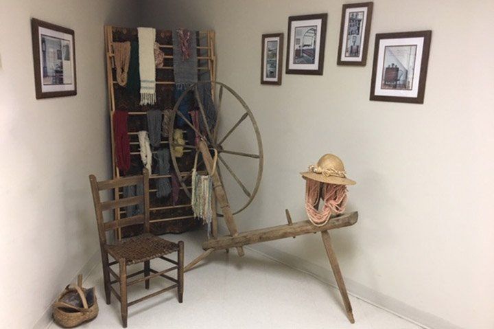 Corner room with spinning wheel, textiles on rack, chair, and framed photos.