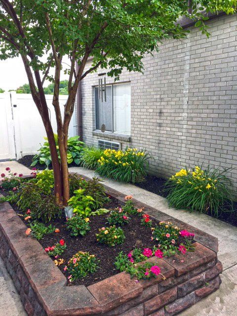 Raised garden bed with colorful flowers and a small tree, next to a brick wall.