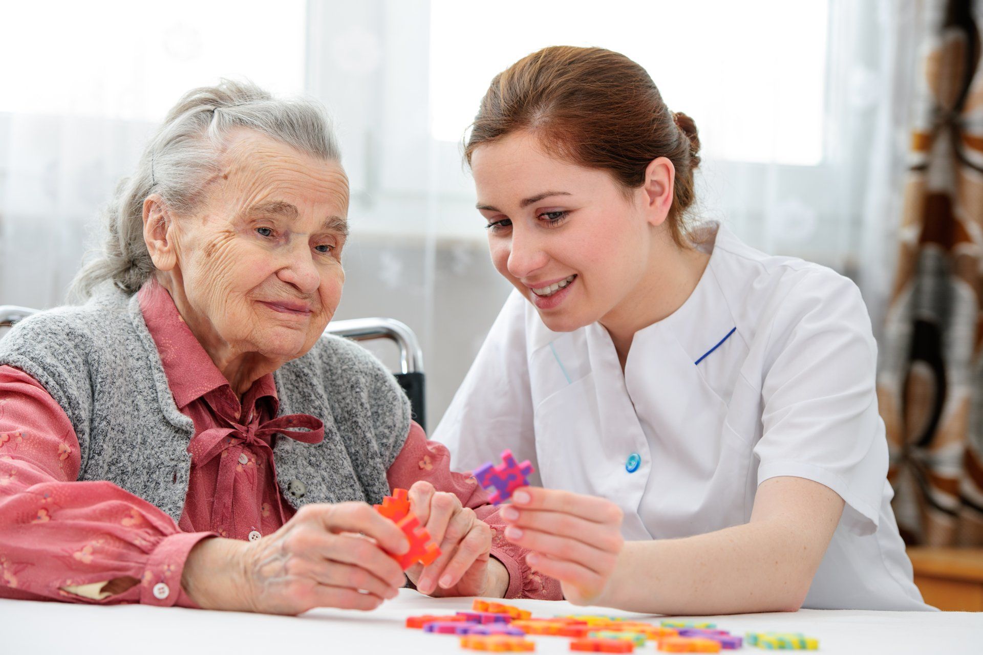 Woman and elderly person playing a puzzle together at a table. The woman smiles.