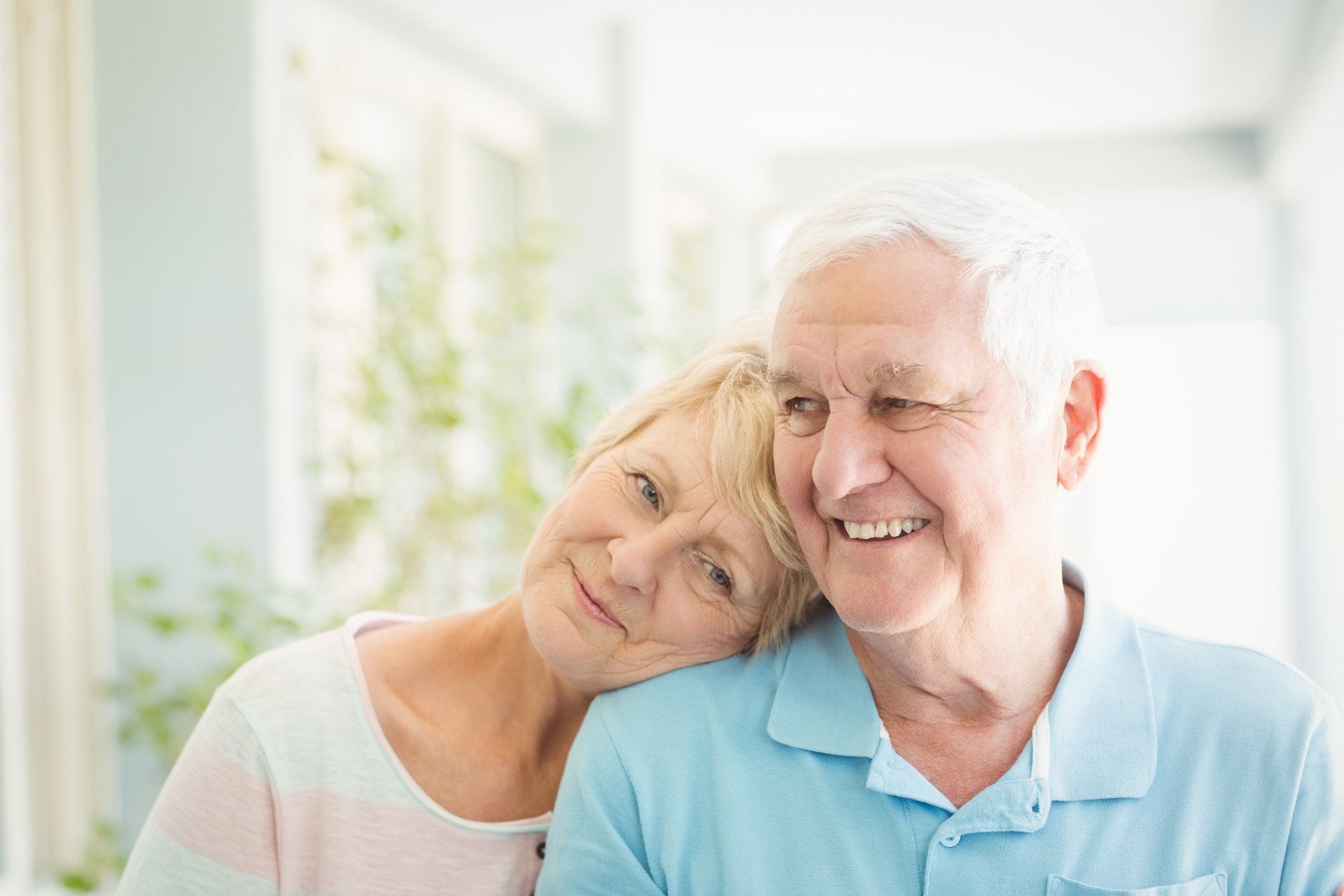 Senior couple smiling; woman resting head on man's shoulder. Natural light, indoors, pastel colors.