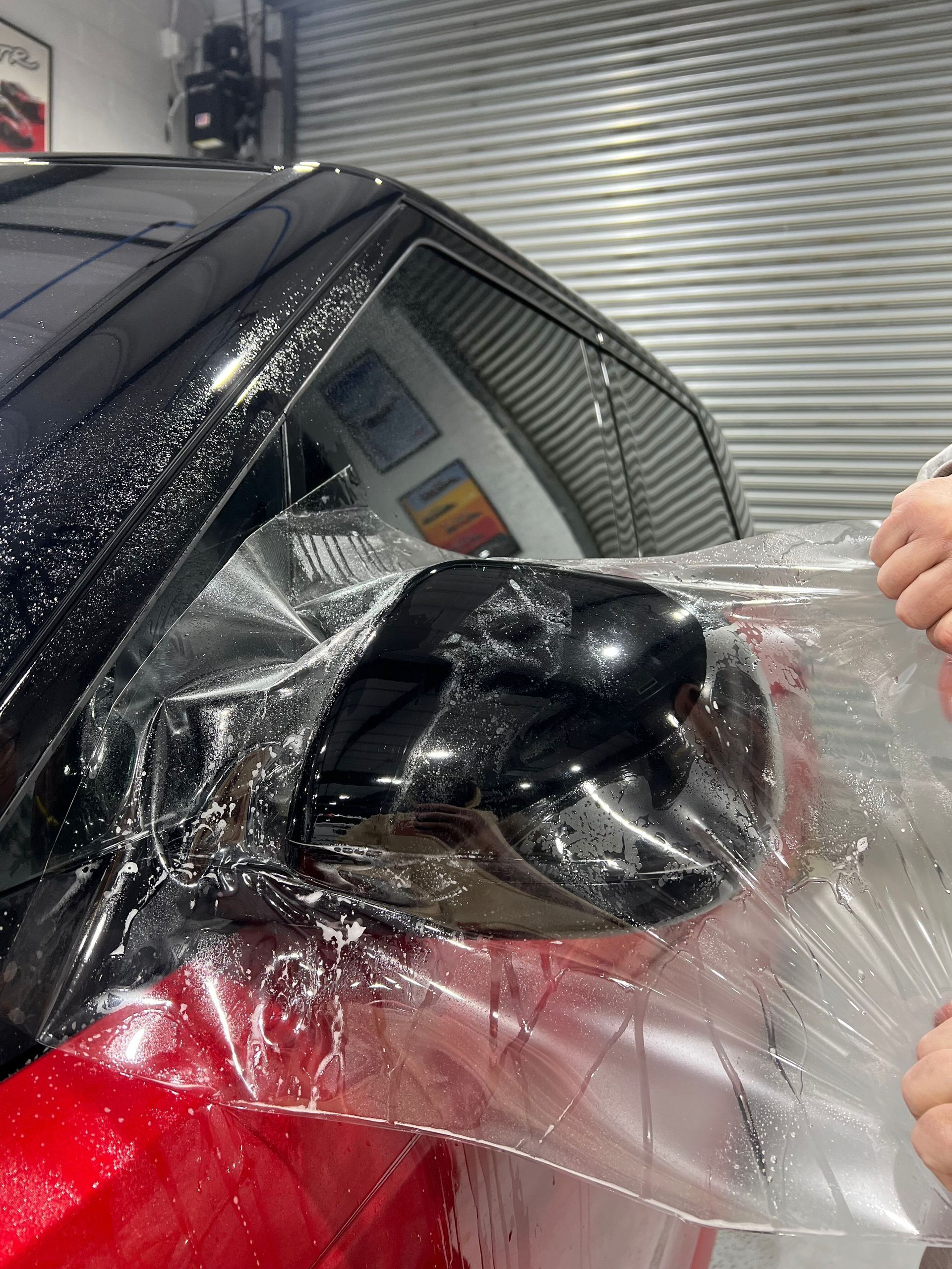 Applying clear film to a black car side mirror. Hands pull the film, in a garage setting.