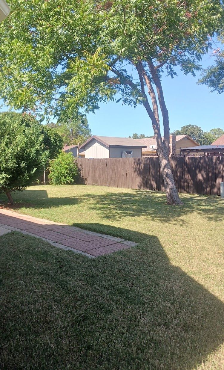 Backyard with a brick patio, green grass, trees, and a wooden fence. A house is visible in the background.