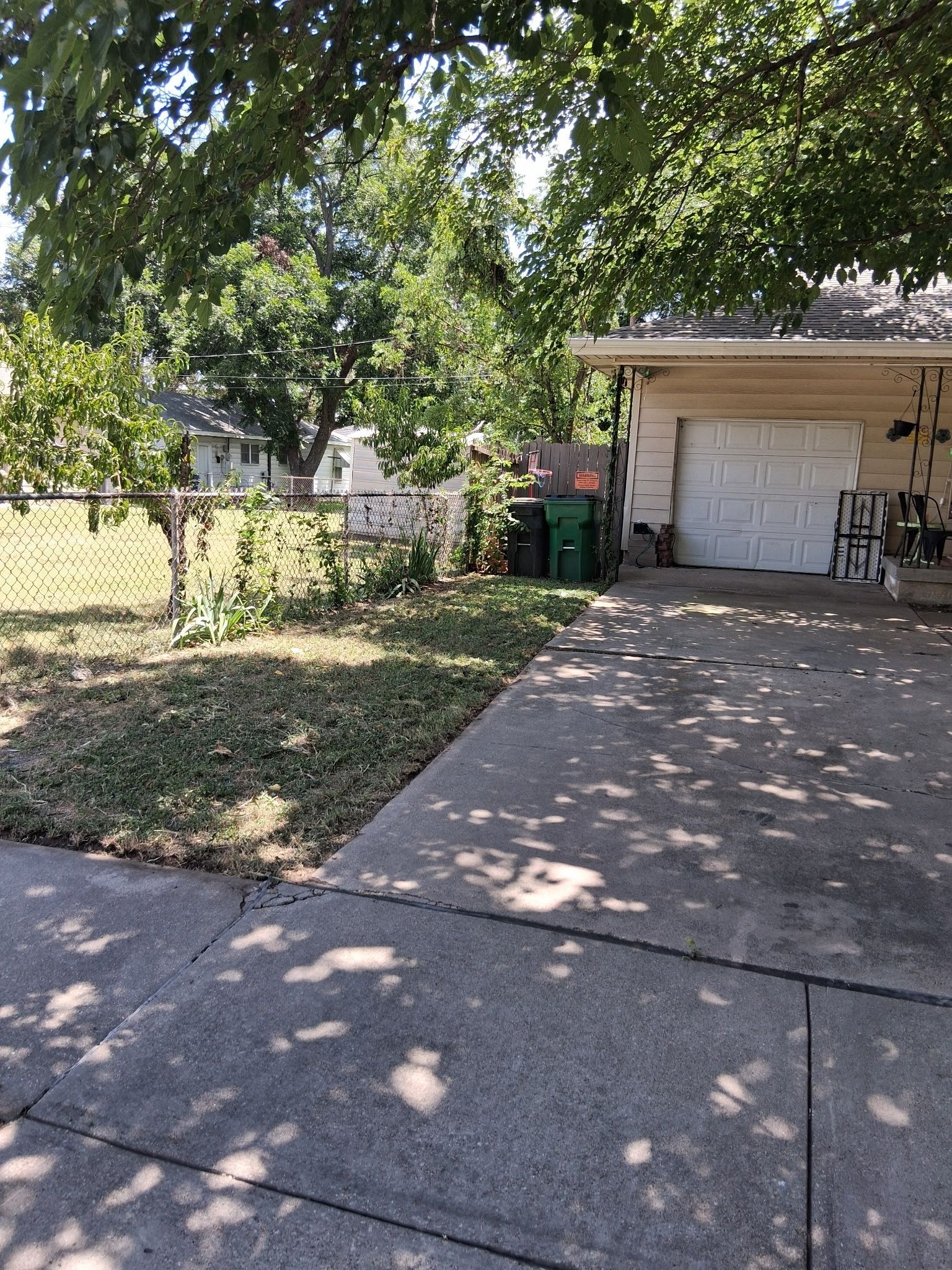 Driveway leading to a garage next to a grassy yard and chain-link fence, under leafy trees.