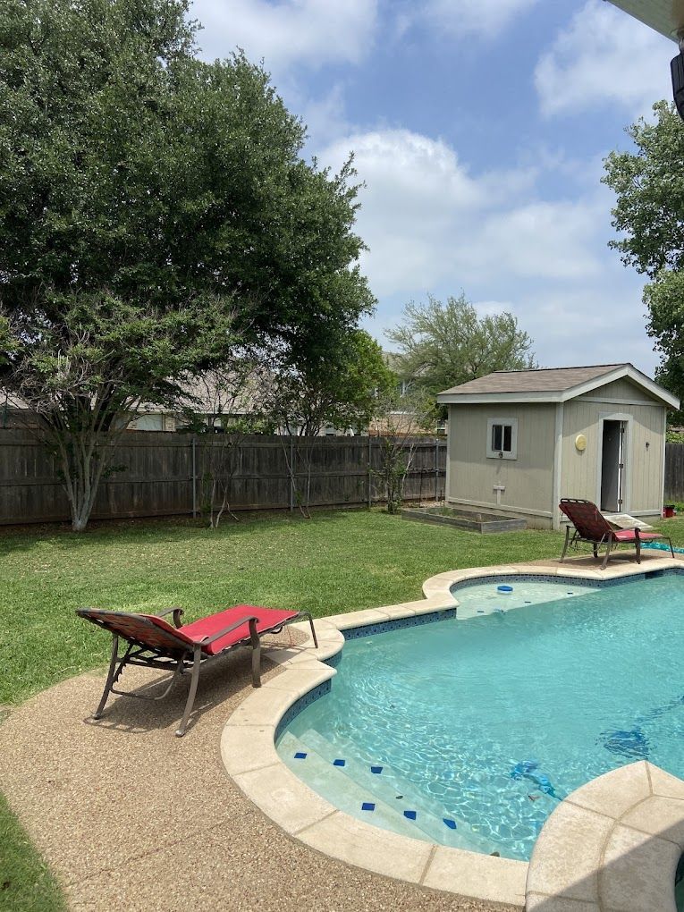 Backyard scene: pool, lounge chairs, shed, tree, and fence under a partly cloudy sky.