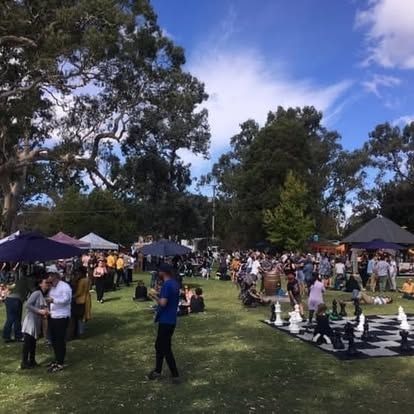 A park festival with a large chessboard. People gather near stalls under trees on a sunny day.