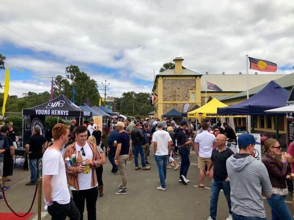 Outdoor market with people milling around stalls under a cloudy sky. An Aboriginal flag flies.