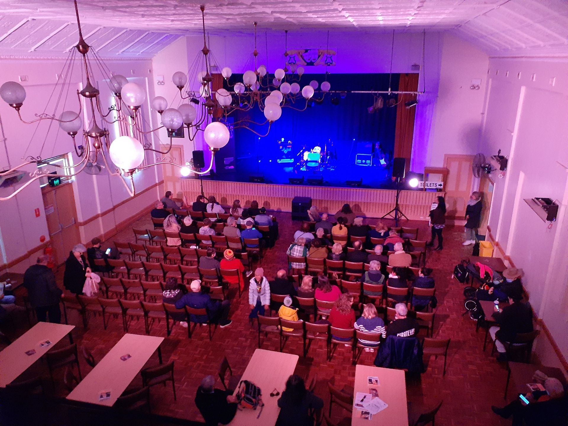 Audience watching a band on stage in an old hall with chandeliers, tables, and rows of chairs.