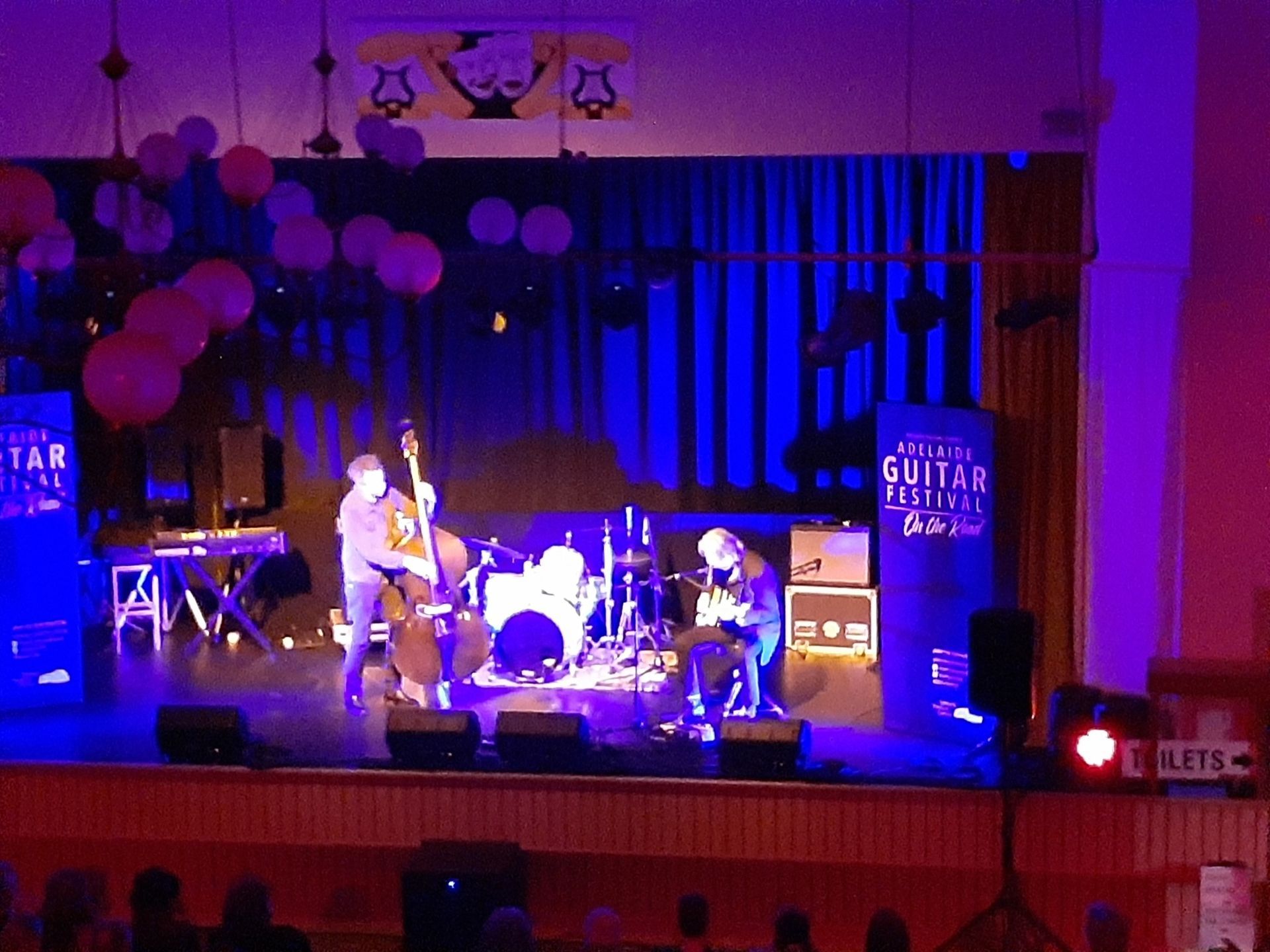 Jazz trio performing on stage, lit by blue and pink lights. Balloons decorate the backdrop.