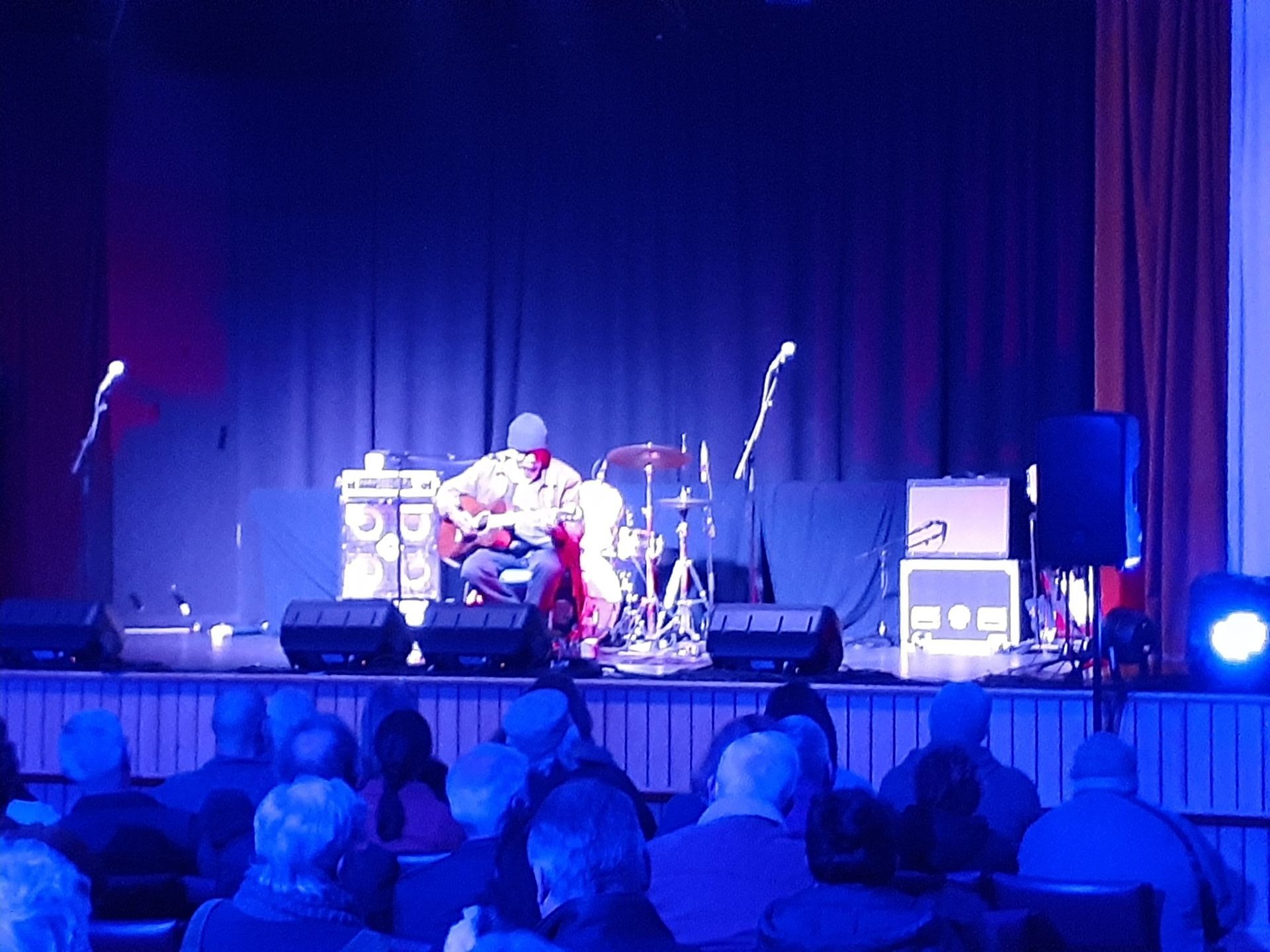 Guitarist performing on stage under blue lights, with an audience in the foreground.