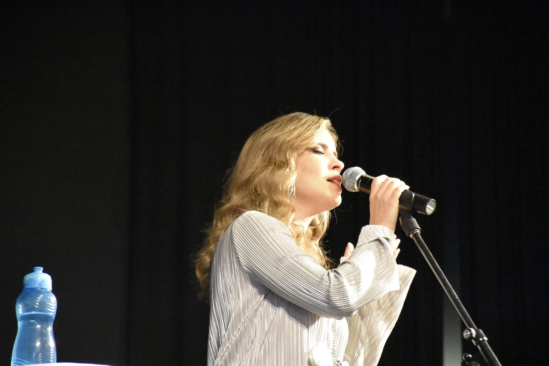 Woman singing into a microphone onstage; wearing a white, textured top. A water bottle sits on a table.