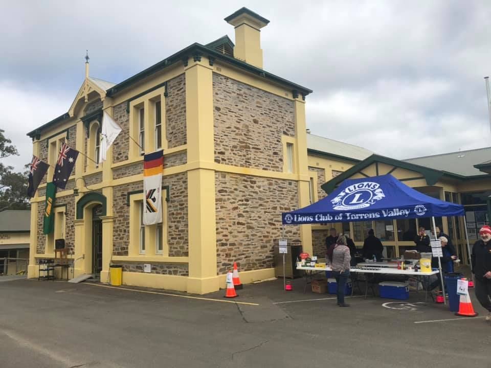 Stone building with Lions Club tent, flags, people; outdoor event.
