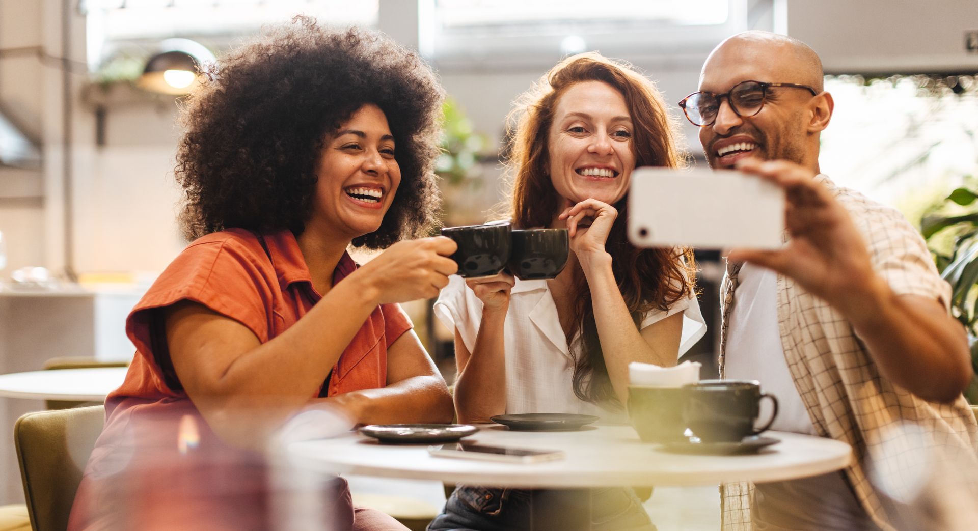 Three people smiling, taking a selfie at a cafe. They hold coffee cups.