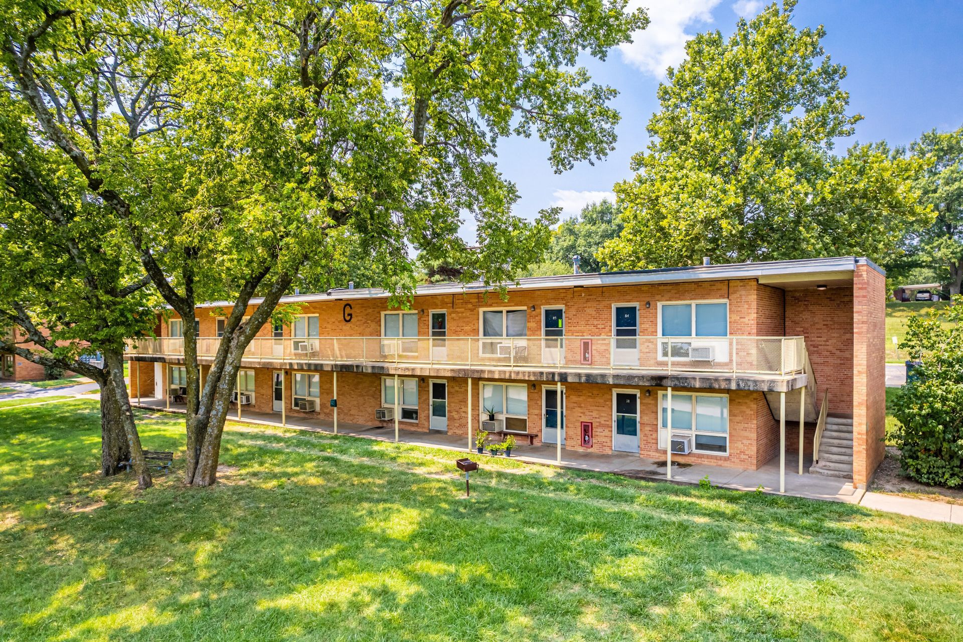 Two-story brick apartment building with balconies, set among trees and green lawn.