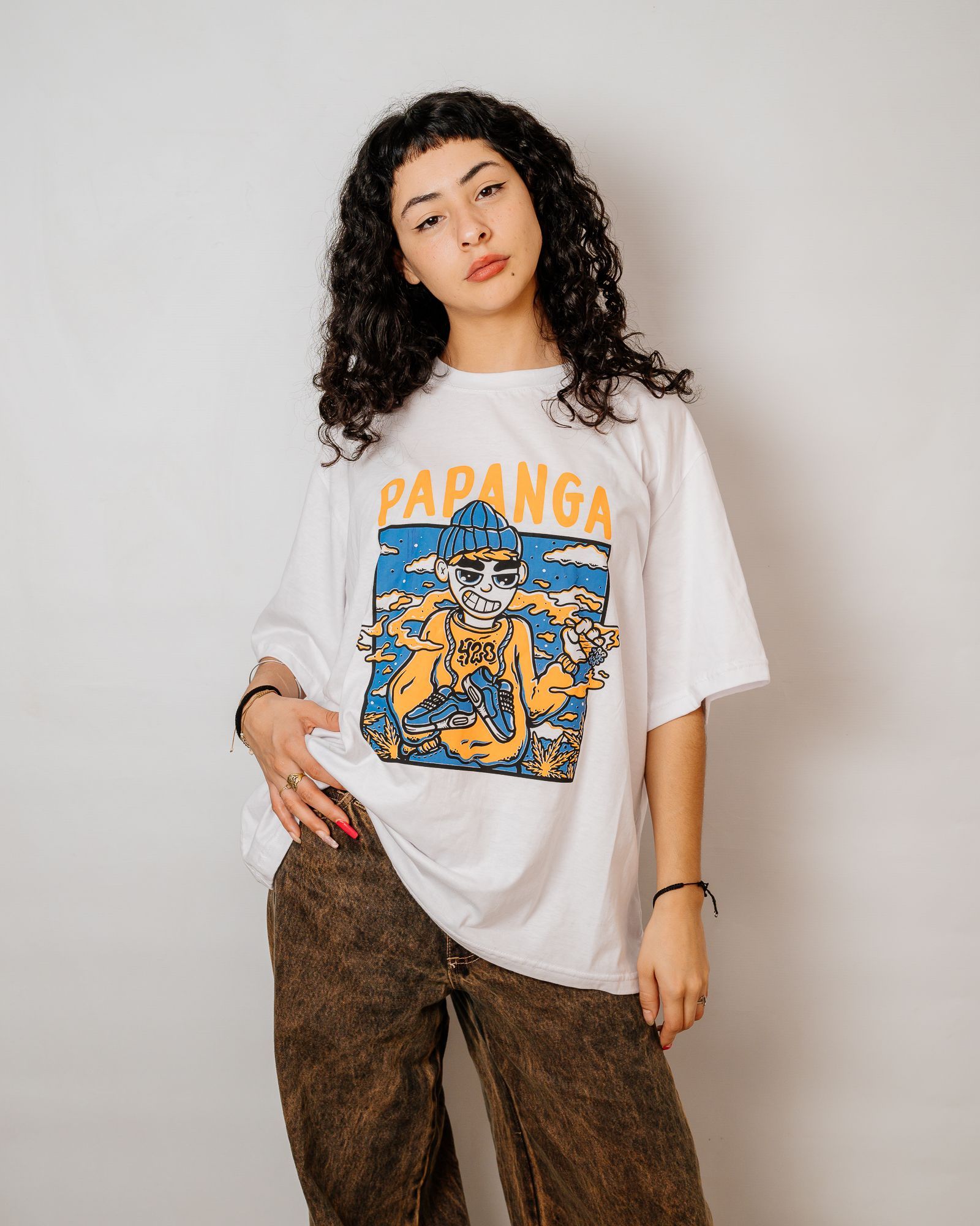 Mujer vestida con camiseta blanca con estampado gráfico, pantalones marrones y cabello oscuro y rizado.