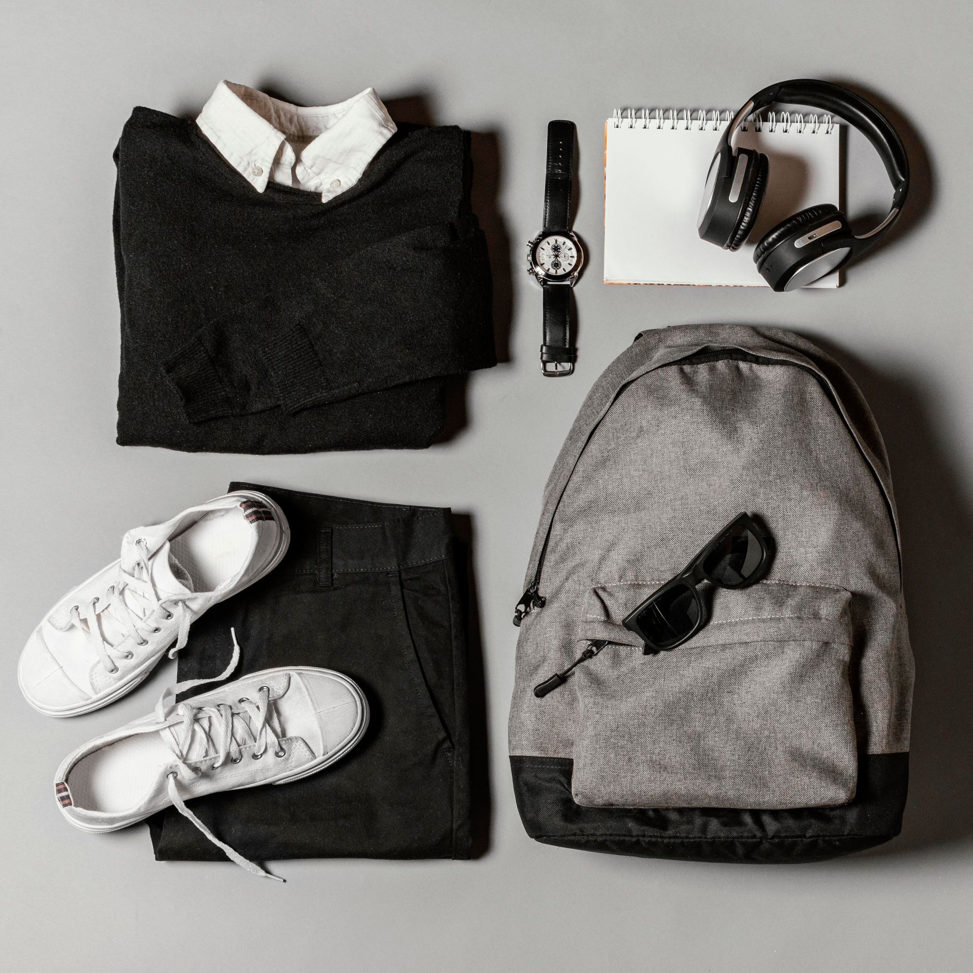 Plano de un conjunto negro con suéter, pantalones, zapatillas, mochila, reloj, auriculares y un bloc de notas.