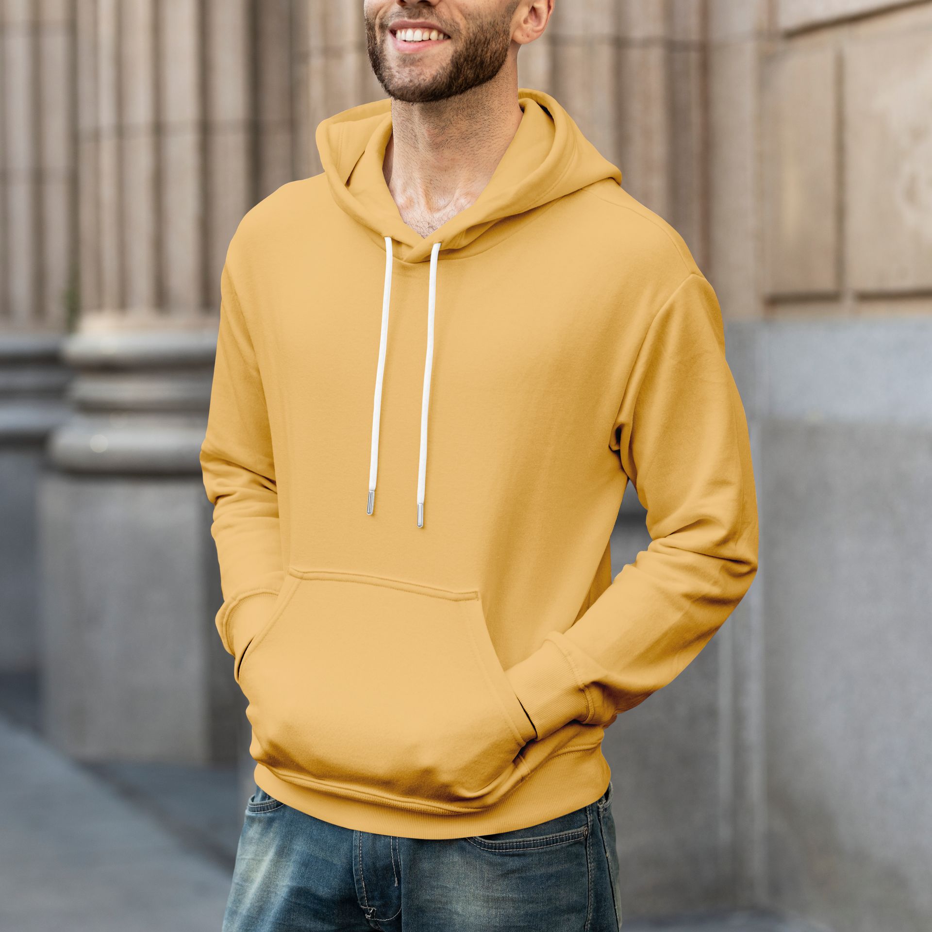 Hombre vestido con una sudadera con capucha de color amarillo mostaza y pantalones vaqueros, de pie afuera.