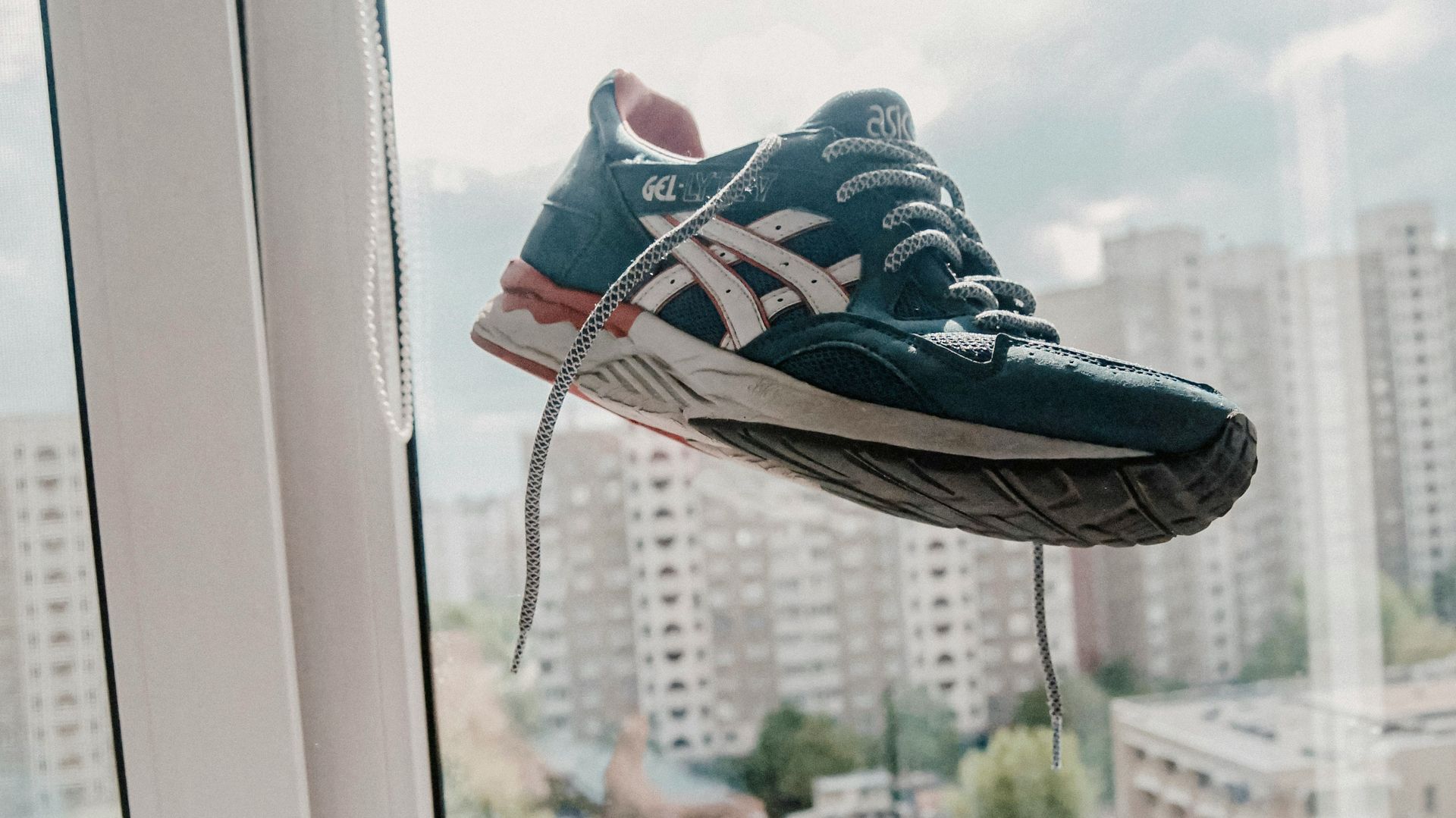 Zapatilla Asics azul y blanca levitando frente a una ventana, con cordones colgando y vista de la ciudad de fondo.