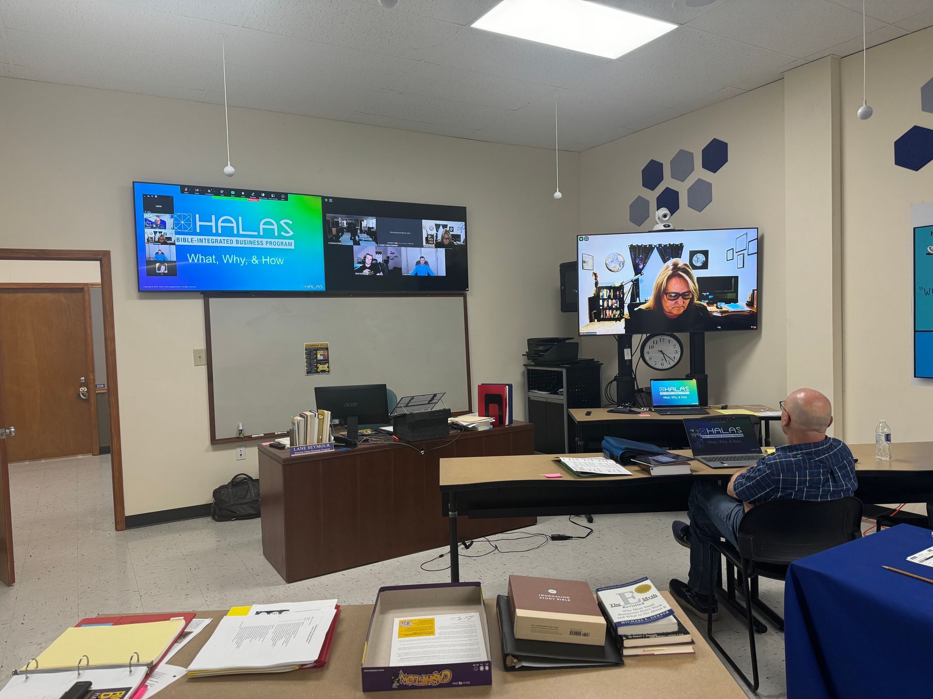 A man is sitting at a desk in a classroom watching a video on a large screen.