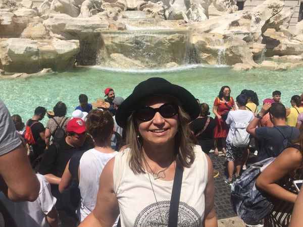 A woman wearing sunglasses and a hat is standing in front of a waterfall.