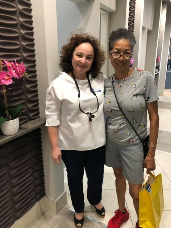 Two women smiling, posing in an office. One in white scrubs, the other in gray with a yellow bag.
