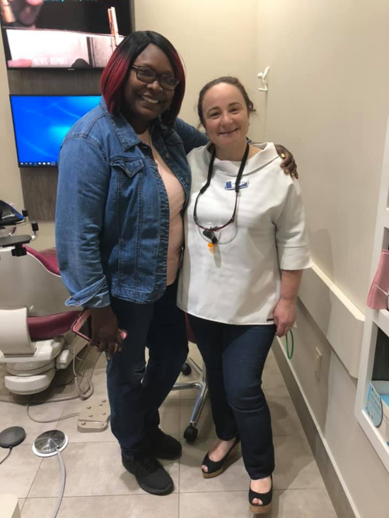 Two women smiling, posing in a dental office. One in a denim jacket, the other wearing a white top and dental tools.