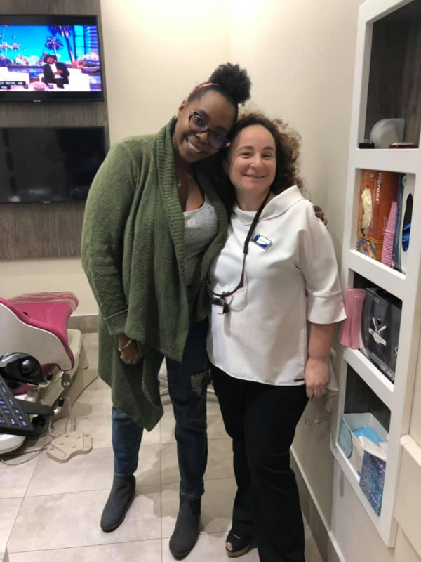 Two smiling women pose, one in a green cardigan, the other in a white top. A dental office is in the background.