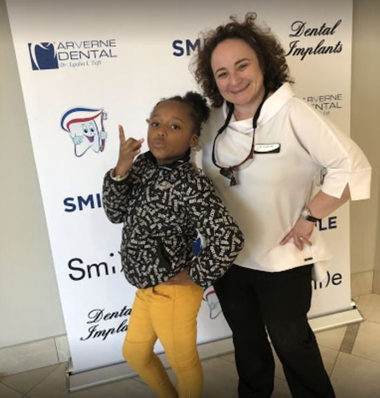 A girl in yellow pants and a woman stand in front of a dental clinic banner.