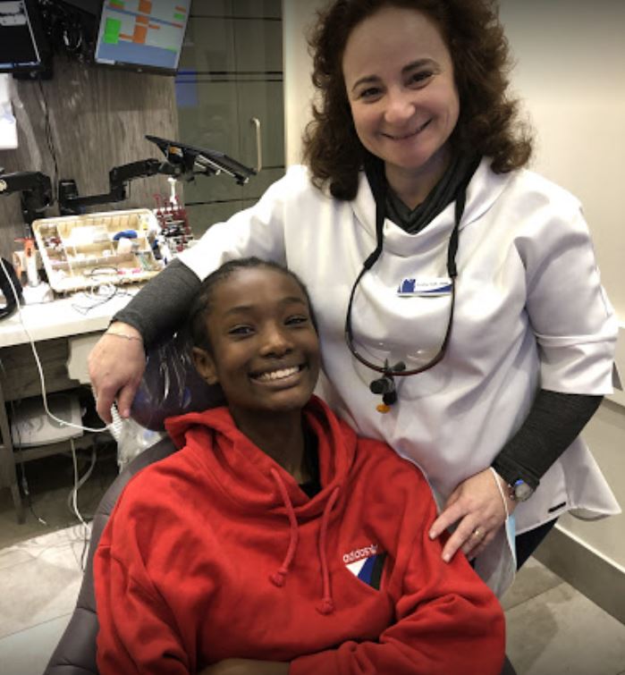 A Black teen in a red hoodie smiles with a dentist in a white coat in a dental office.