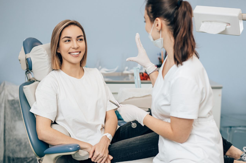 Woman smiles at a dentist in a dental chair; the dentist gestures with gloves in a clinic.
