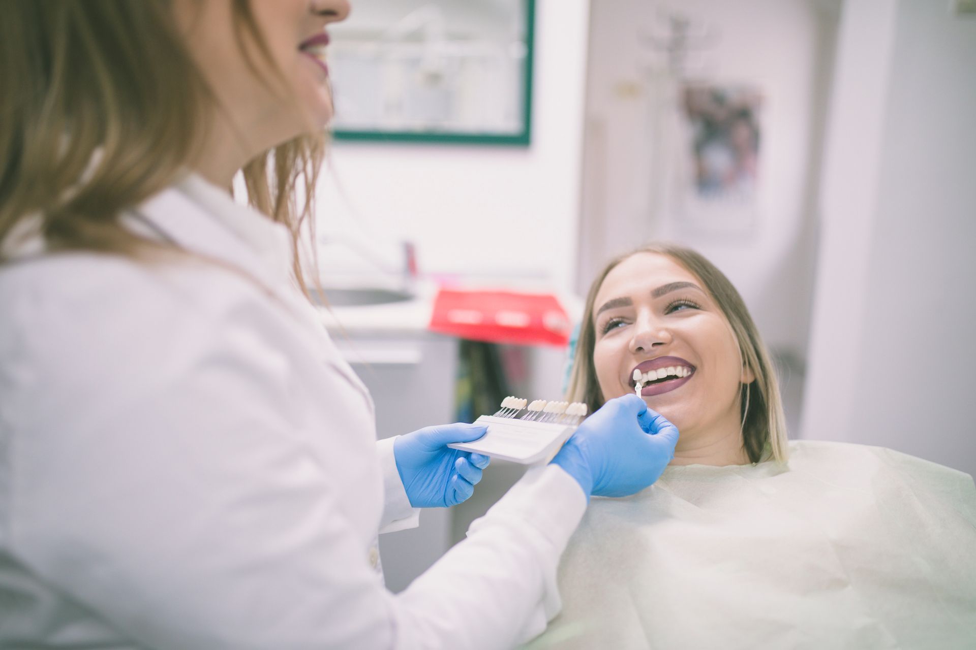 Dentist examining a smiling woman's teeth with a shade guide in a dental office.