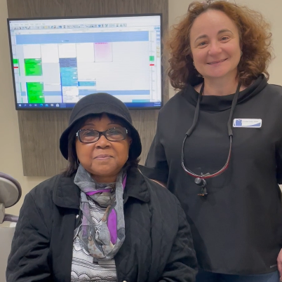 Woman in hat and dental worker smiling in a dental office. Schedule displayed behind them.