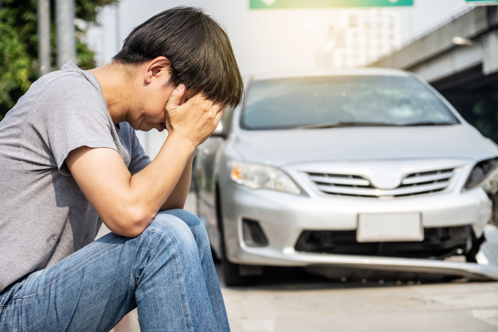 A man in jeans and a gray shirt sits with his head in his hands in distress next to a damaged silver car on a road.