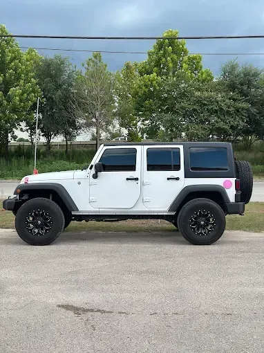 White Jeep Wrangler with black wheels, parked on asphalt with trees in the background.