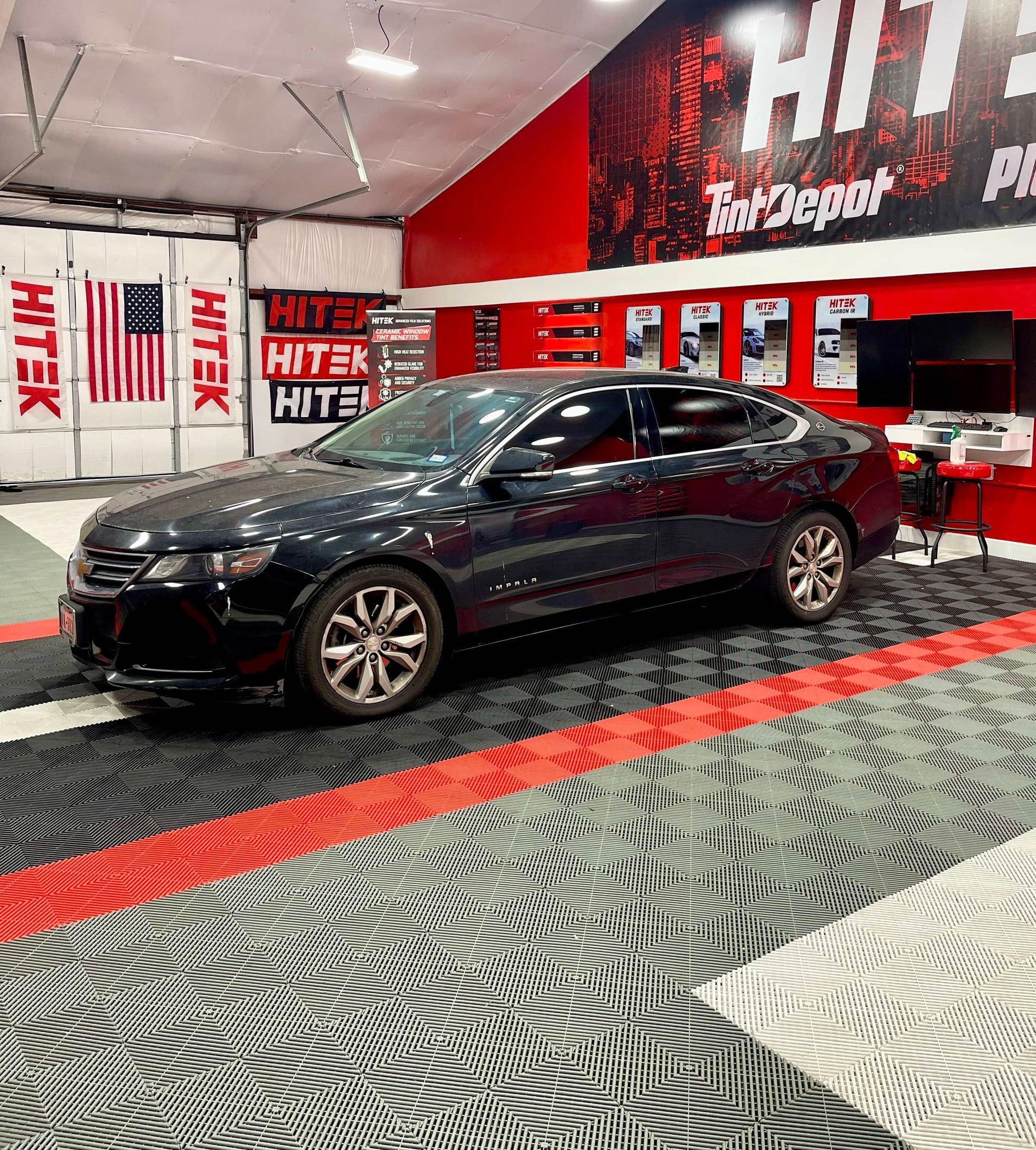 Black sedan parked inside a garage with red and black floor tiles. American flag in the background.