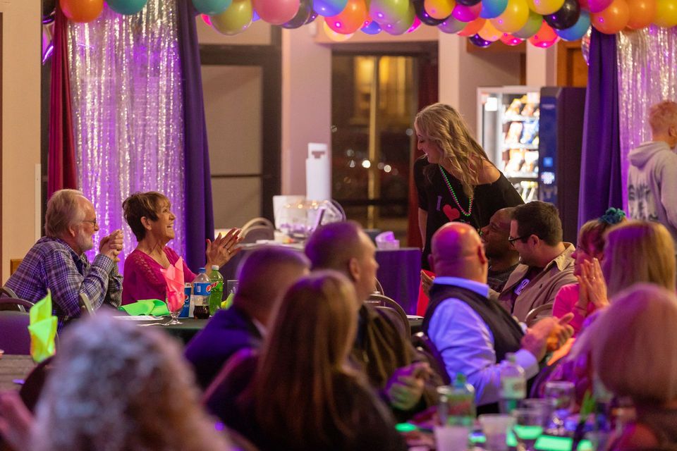 A group of people are sitting at tables at a party with balloons hanging from the ceiling.