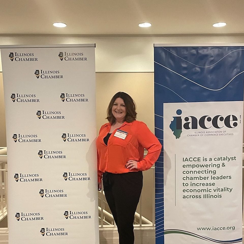 A woman in an orange shirt is standing in front of a banner for the illinois chamber of commerce.