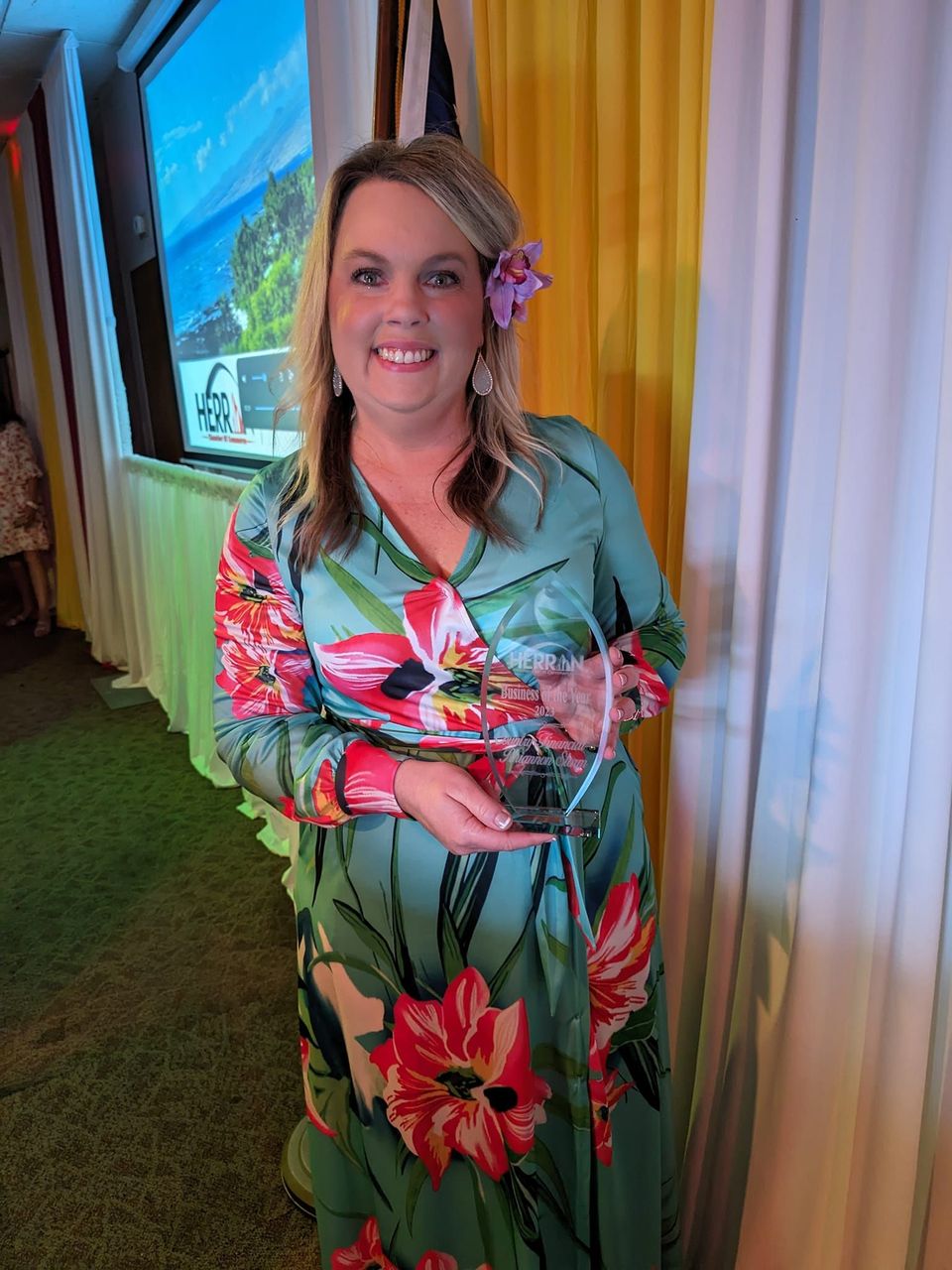 A woman in a floral dress is holding a trophy and smiling.