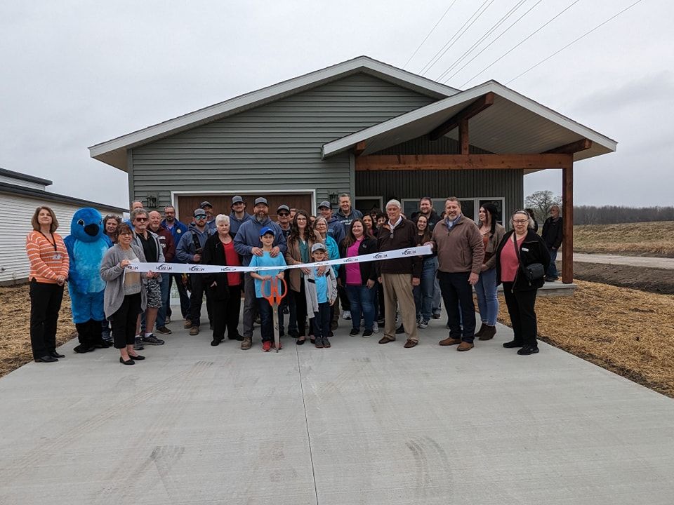 A group of people are standing in front of a house holding a ribbon.