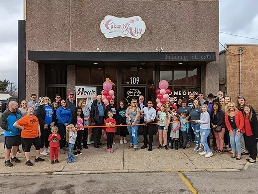 A large group of people are standing in front of a building cutting a red ribbon.