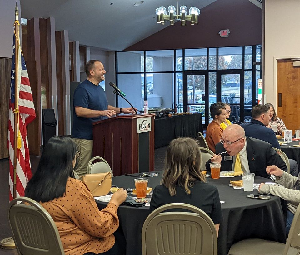 A man is standing at a podium giving a speech to a group of people sitting at tables.