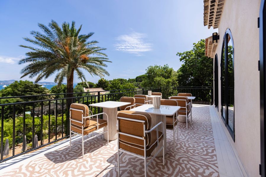 An outdoor terrace with white-framed chairs and tables, a peach umbrella, and a patterned rug against a light wall.