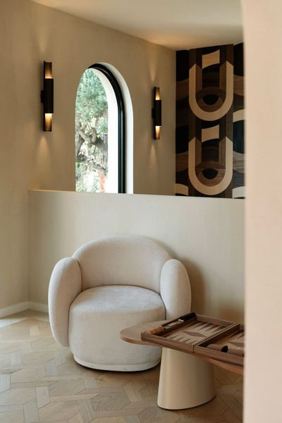 A white textured armchair sits beside a wood backgammon board in a beige room with a dark geometric wall and arched window.