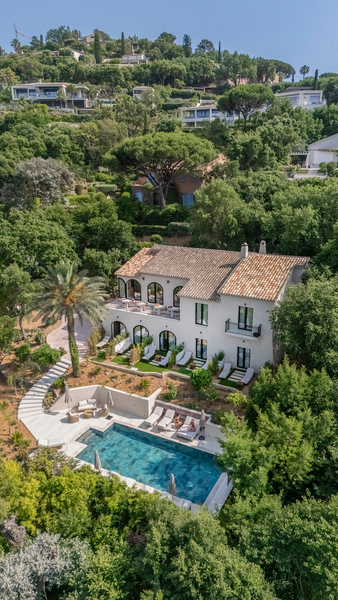 A white villa with a terracotta roof nestled in lush hills, featuring a swimming pool and sun loungers on a stone patio.